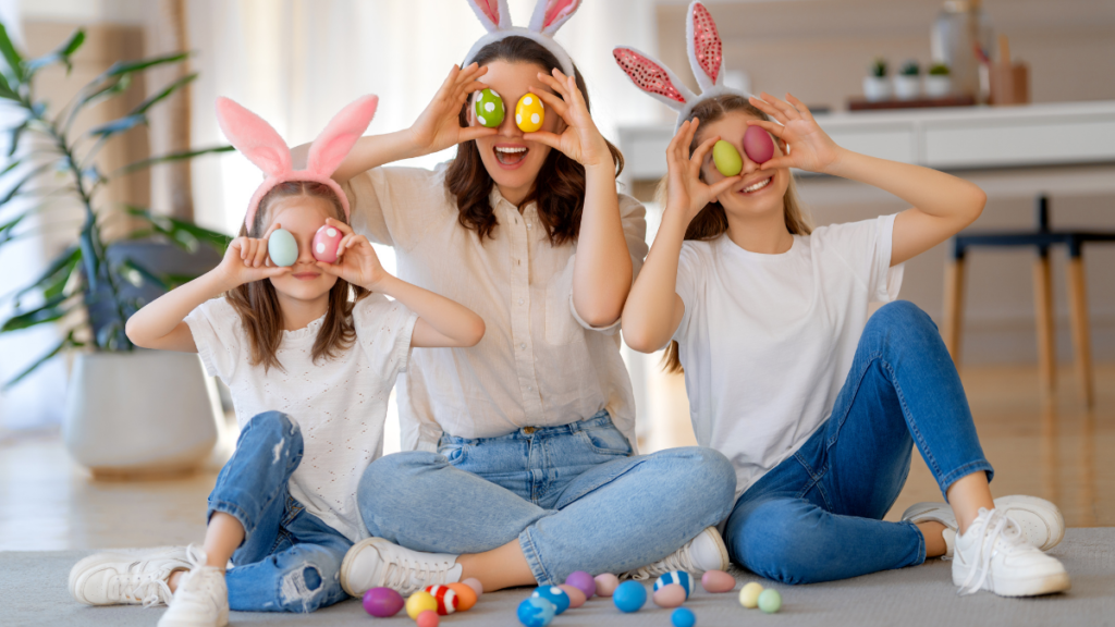 Mãe e duas filhas crianças, vestindo blusa branca e calça jeans, orelhinhas de coelho de Páscoa, e segurando ovos de Páscoa coloridos na frente dos olhos, sorrindo. Tríduo Pascal em Casa.